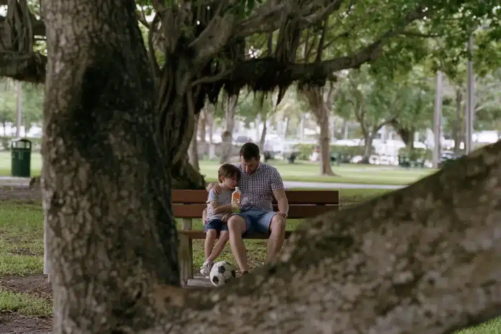 A father and his young son sitting together on a bench under banyan shade in Holiday Park, Fort Lauderdale