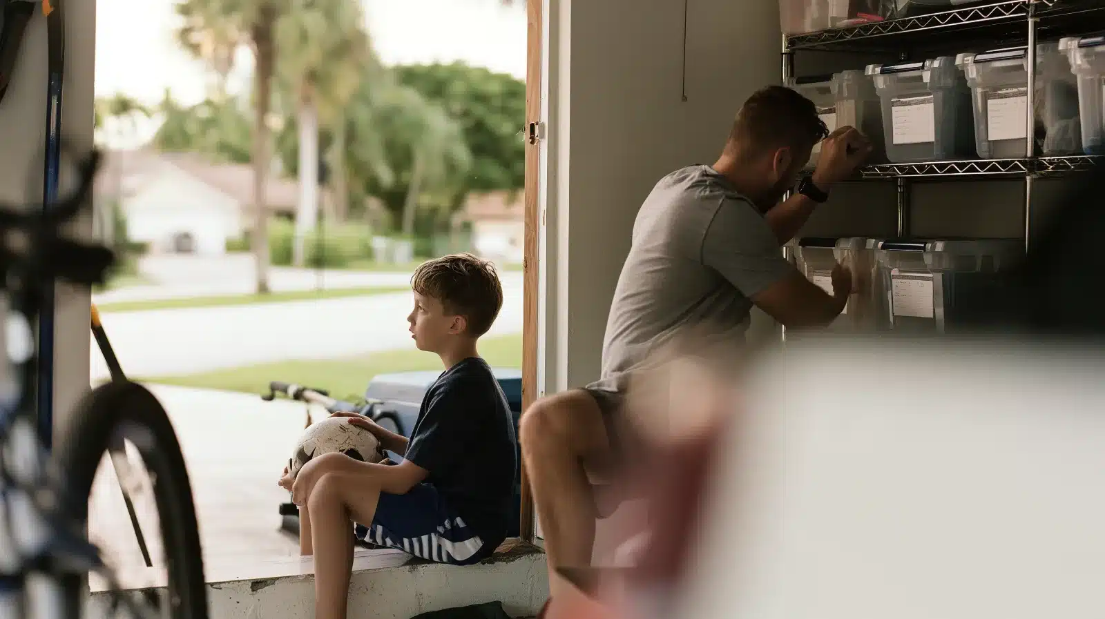 Boy holding a soccer ball watching his exacting father label bins in the garage, an illustration of the childhood unrelenting standards behind OCPD