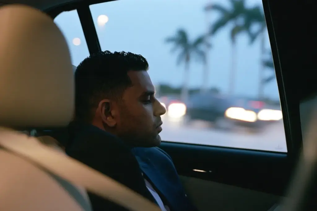 A South Asian man in the back seat of a car on I-95 at dusk, looking out the window
