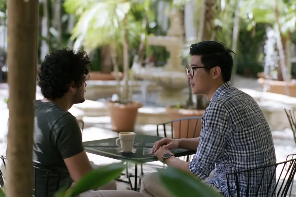 Two men in their thirties talking at a café table in a shaded Fort Lauderdale courtyard