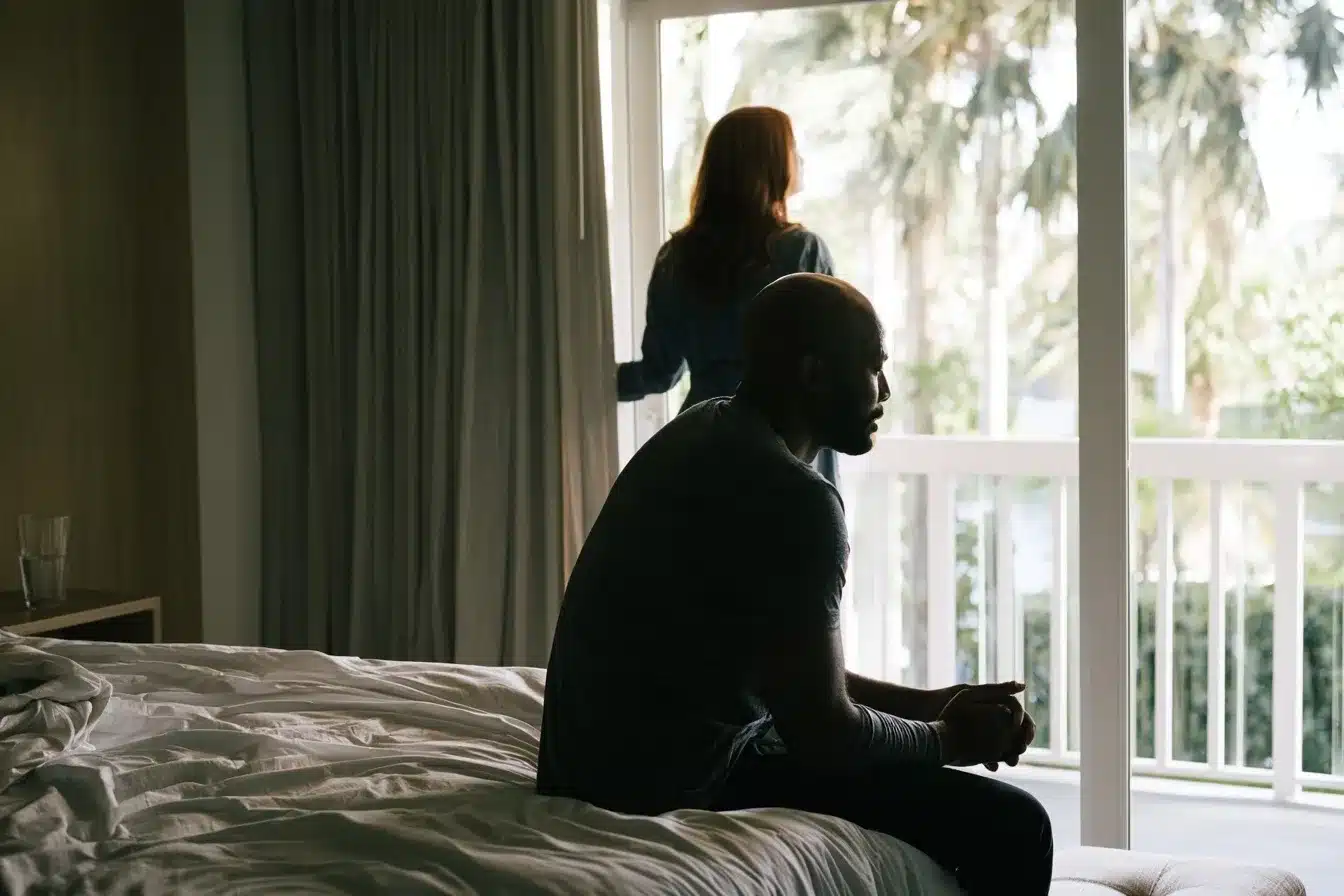 Couple in a Boca Raton bedroom after tension, she stands at the sliding glass door while he sits on the edge of the bed